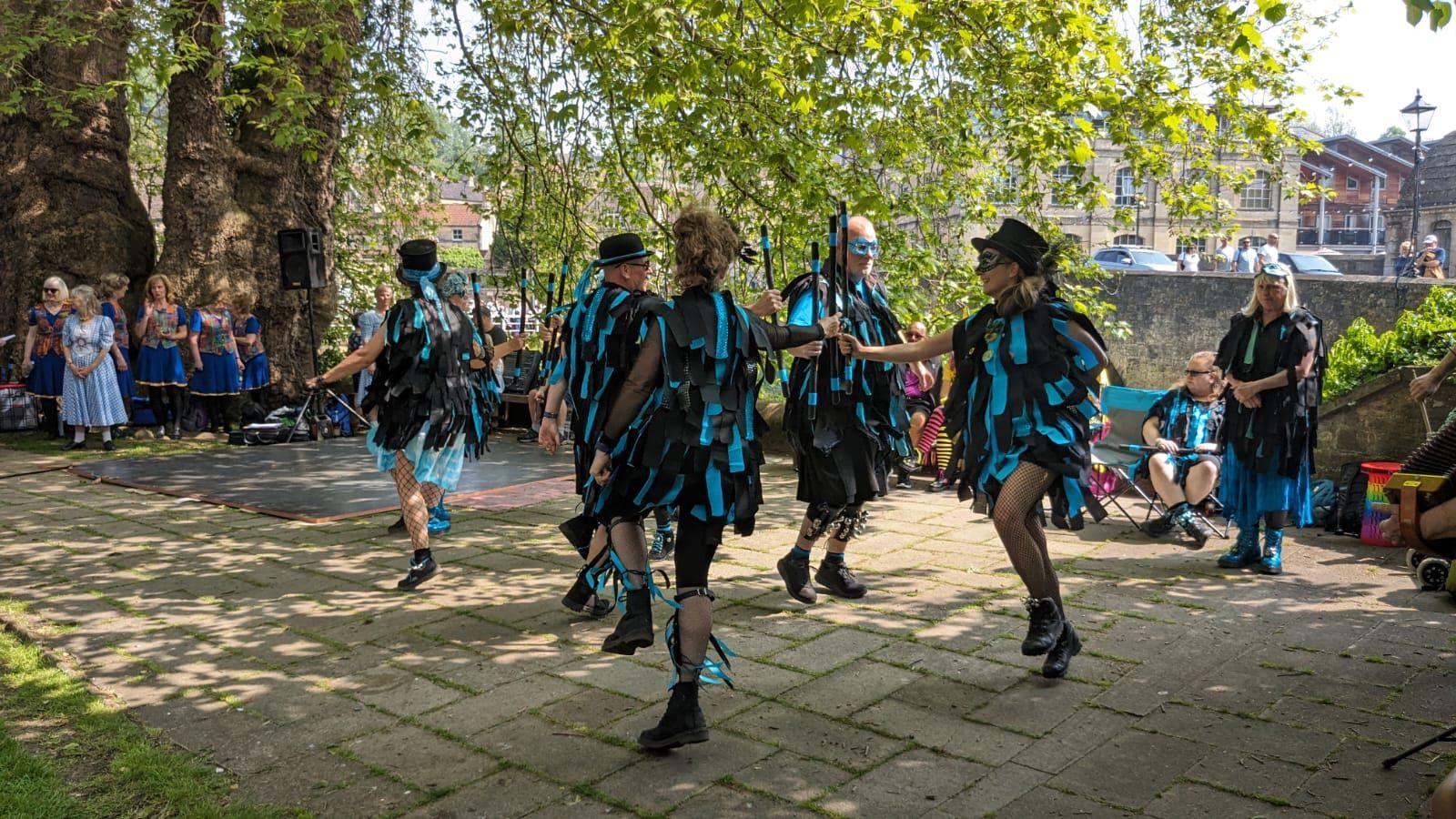 Several people wearing black and blue matching outfits, eye masks, and some with black hats, dancing on a brick path with a large tree in the background. There are some people watching , including a group of women in blue dresses.