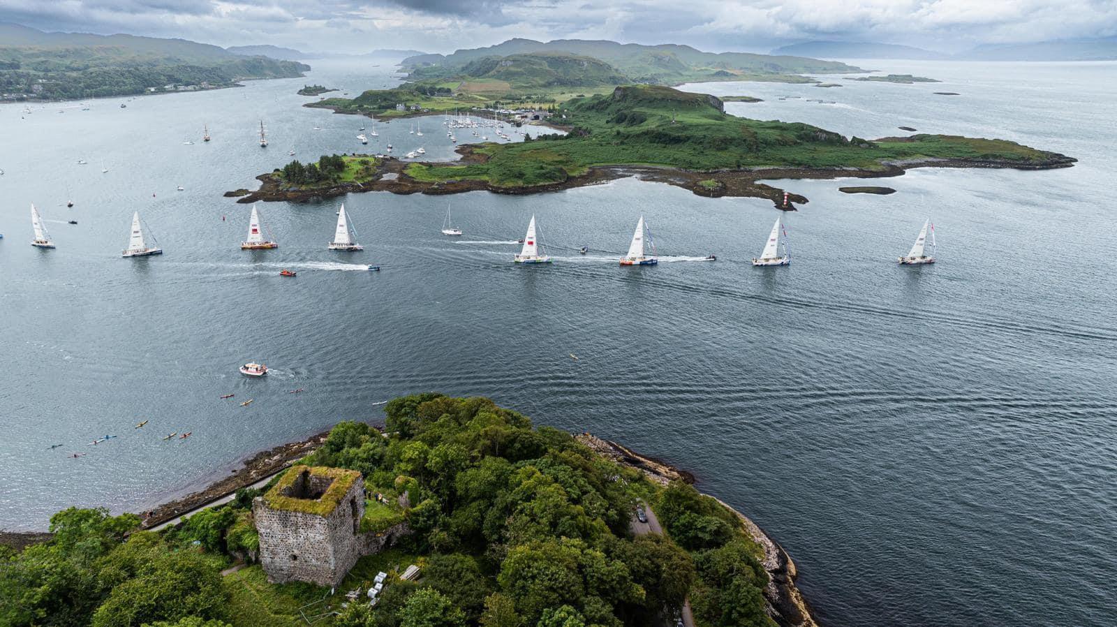 A panoramic view of eight boats sailing on the sea past two big green verges