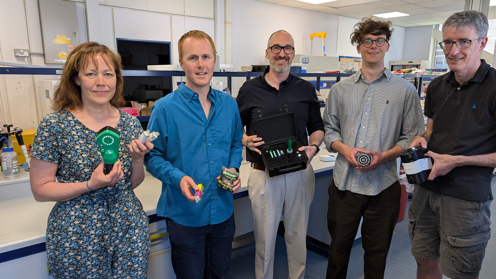 Professor Christopher Pudney is standing with four other members of the Team Harm Reduction, one is a woman, one if a young man and the other two are older men. Each one of the team is holding a device and showing it to the camera. They are standing in a laboratory next to work desks.