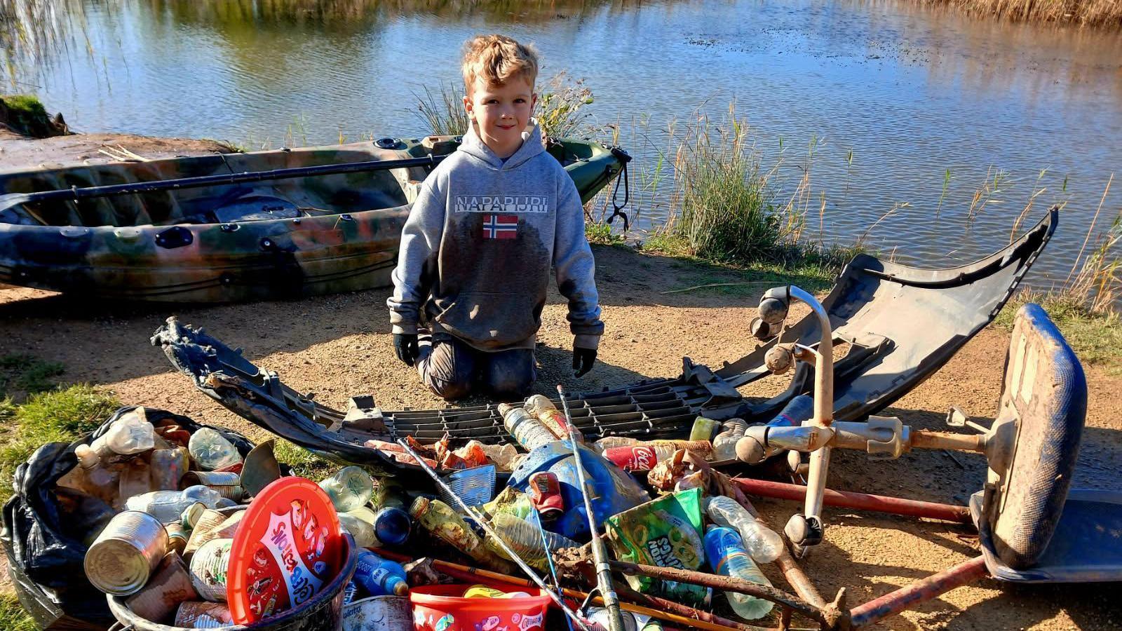 Logan Currie, 7, kneeling in front of a pile of rubbish he and his father collected from a pond. There are bags of cans and a car bumper. He is kneeling next to a camouflaged kayak which his dad used to collect floating rubbish.
