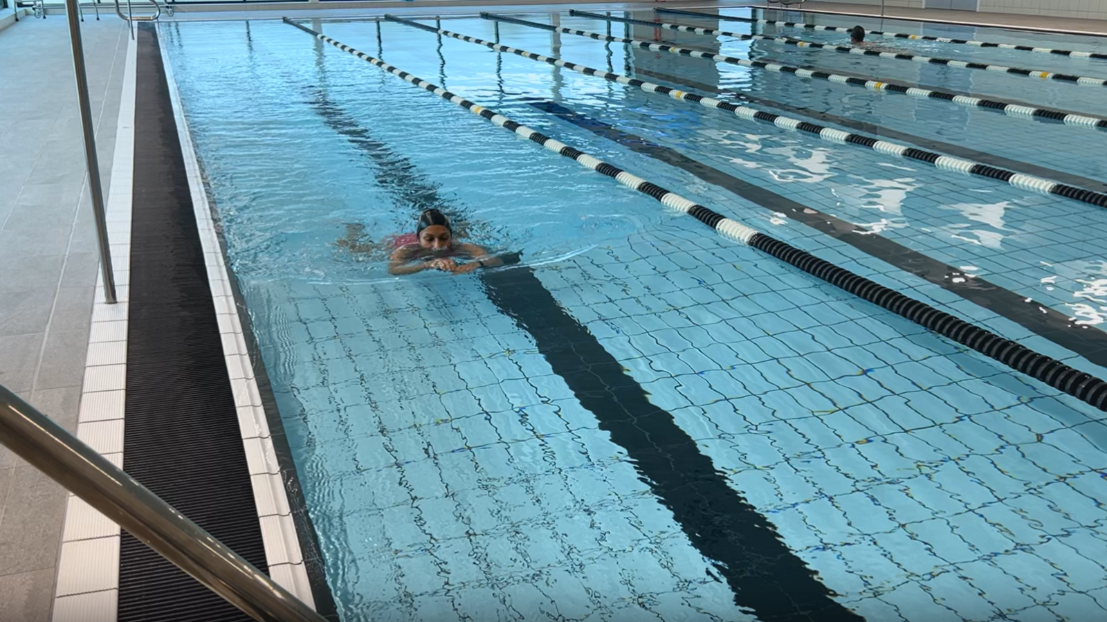 A woman wearing a pink swimming costume and a black swimming hat swimming a length of an indoor swimming pool.