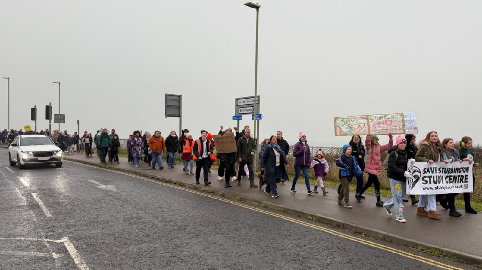 Protesters march along the pavement alongside a main road holding banners. They are made up of adults, children, and some dogs.
