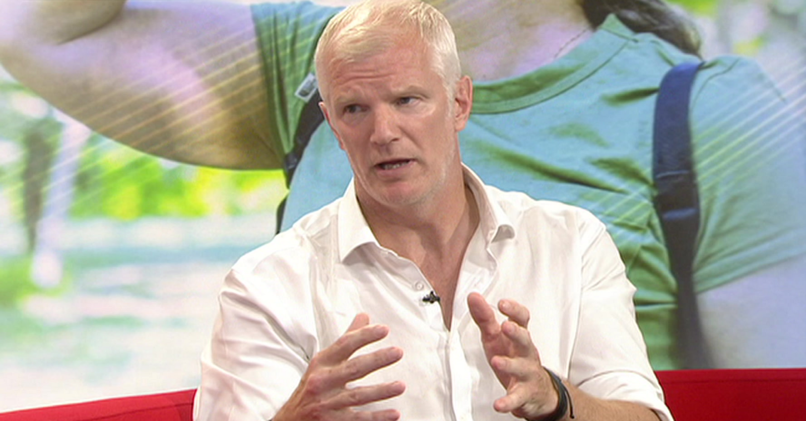A man with short white hair is wearing an open-neck white shirt and sitting on a red sofa in a TV studio. There is a photographic backdrop behind him illustrating the heatwave, with a woman in summer clothes and a backpack.