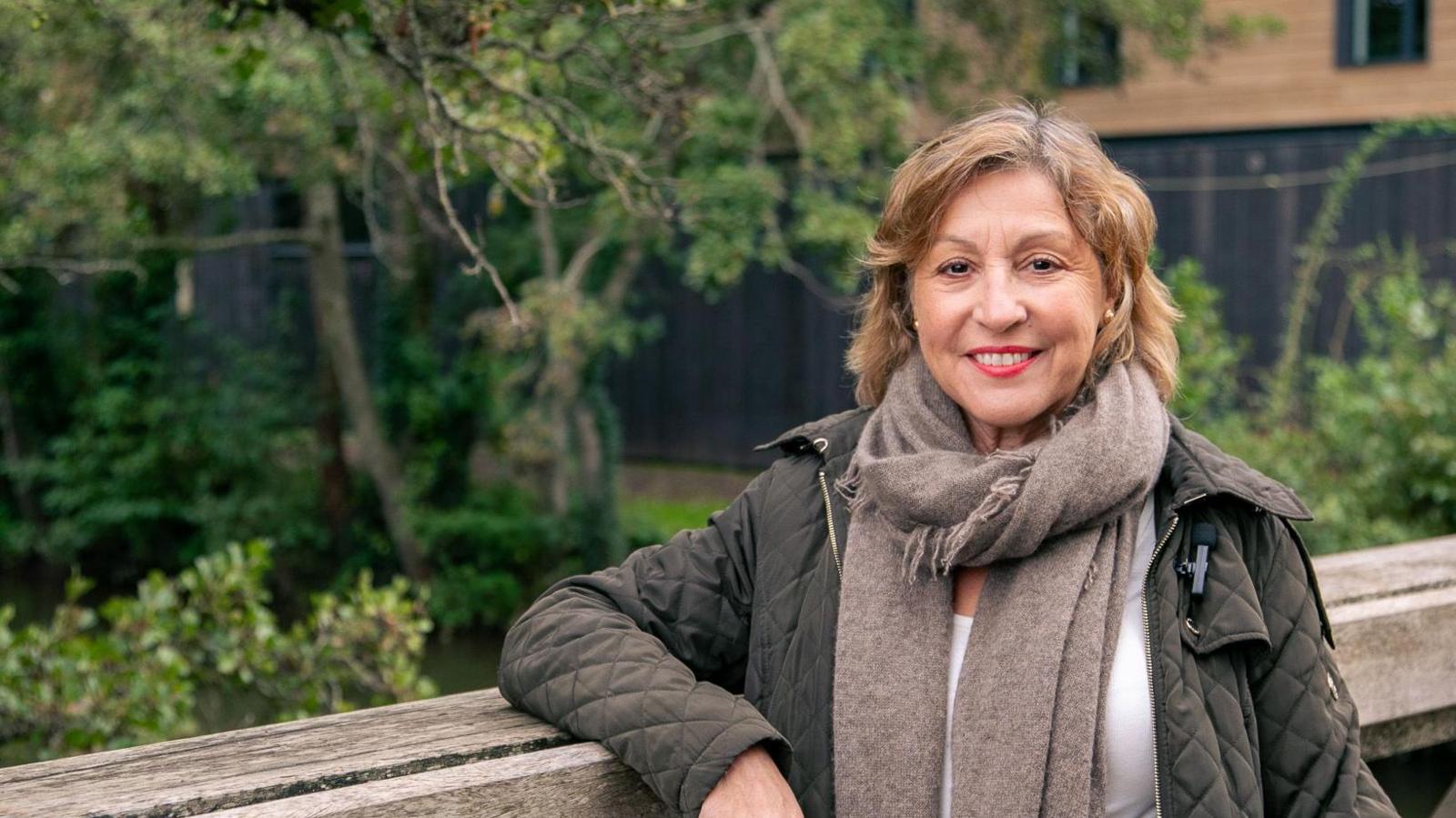 Rebecca Pow in a quilted coat leaning on a fence by a tree