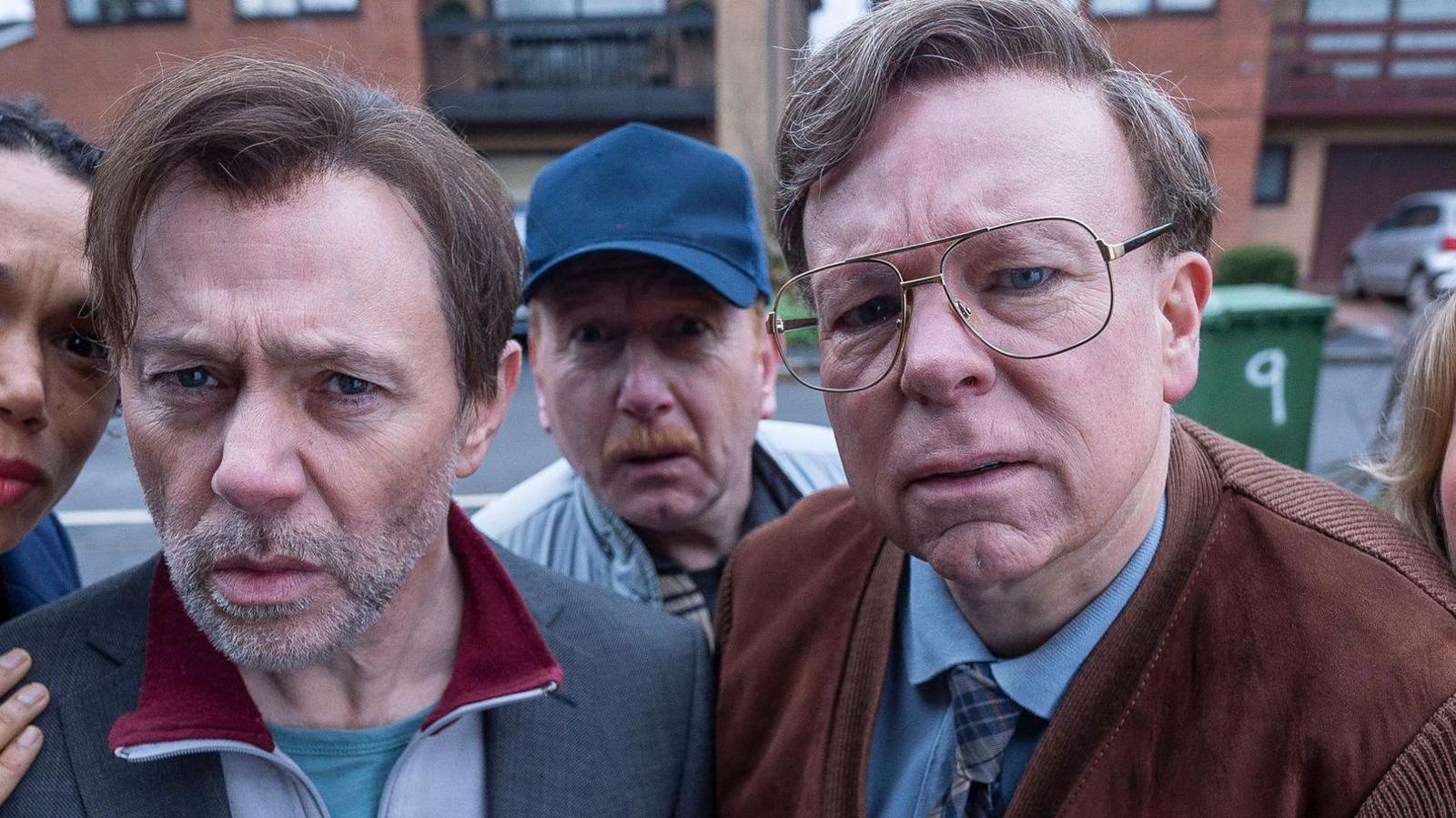 Actors pose in character, with heads and shoulders seen. A group of five are huddled in front of what seems to be a door cam and are looking at it quizzically. Two men are at the front. The one on the left has brown hair that is long on top and grey stubble. He is wearing a grey suit jacket and tracksuit top. The one on the right has seventies-style gold glasses, a brown suede jacket, blue shirt and plaid tie.
