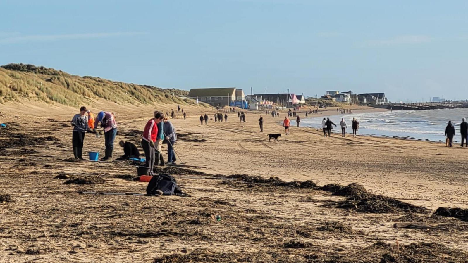 People on a sandy beach at Camber clearing up debris with the sea and buildings in the background.