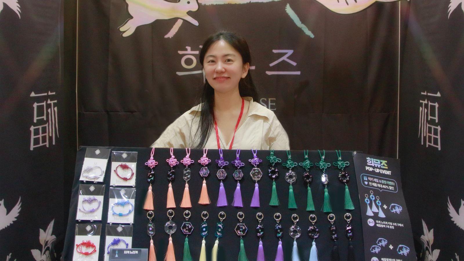 Choi Nyun-hee smiles at the camera, wearing a yellow collared shirt. She has long black hair and a red lanyard. Perched in front of her is a black board with keychains and bracelets hanging on it.