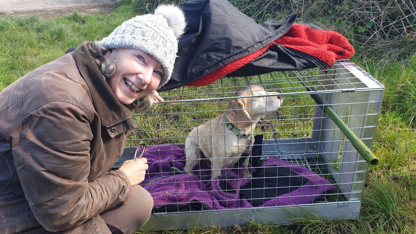 Maxine Young is crouching down to a crate which has a small pale labrador puppy in it. Ms Young is smiling and wearing a brown coat and woolly hat. The puppy is sitting on a fleece in the crate which is lying on grass.