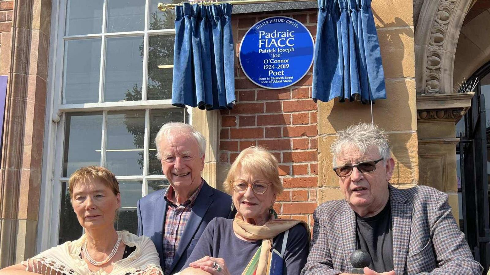 A circular blue plaque with large white writing that reads - Ulster History Circle Padraic Fiacc 1924-2019 - is unveiled on a red brick wall, with dark blue curtains pulled back on each side of it. Below it are four people, two men and two women. The men have grey hair and the women have blonde hair.