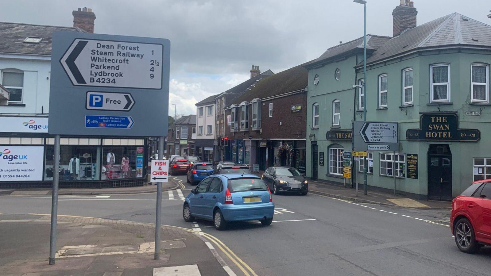 Lydney high street is pictured with cars driving in both directions on the road. Beside the road is the swan hotel, road signs and a charity shop.