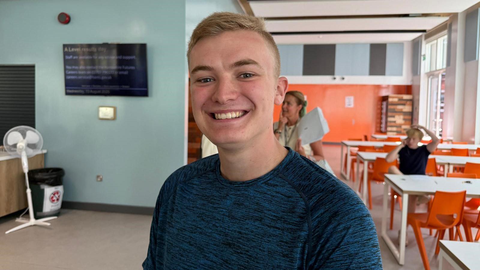 Dante Thompson is smiling at the camera. He has short blonde hair and wears a dark blue marbled print t shirt. In the background behind him there are some white canteen tables with orange chairs and a young man sat down talking on the phone.