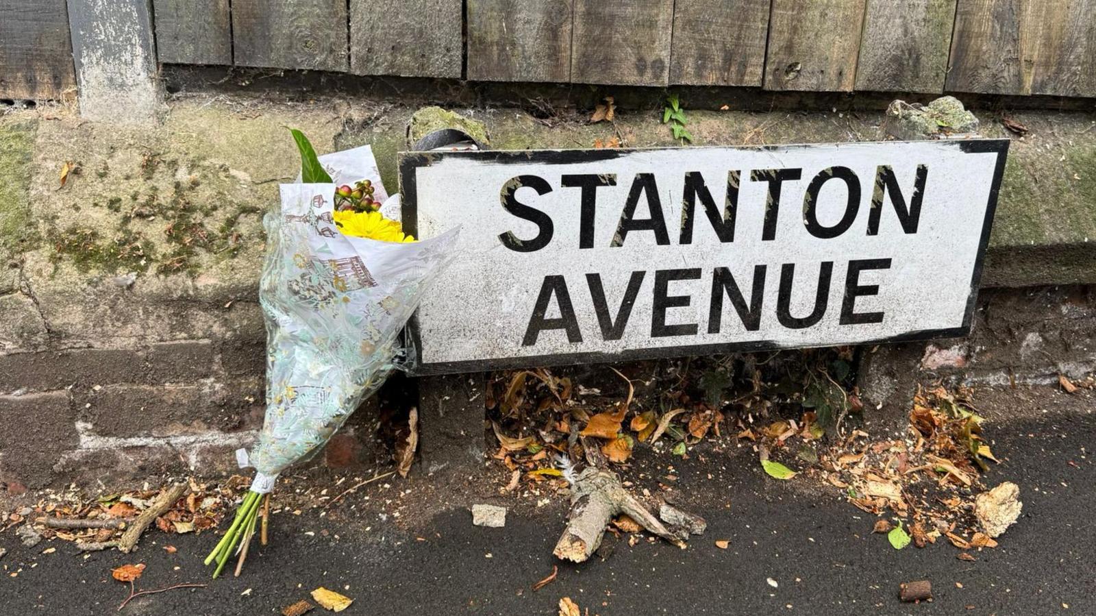 A bouquet of flowers in wrapping leans on the road sign for Stanton Avenue, with leaves and twigs around it