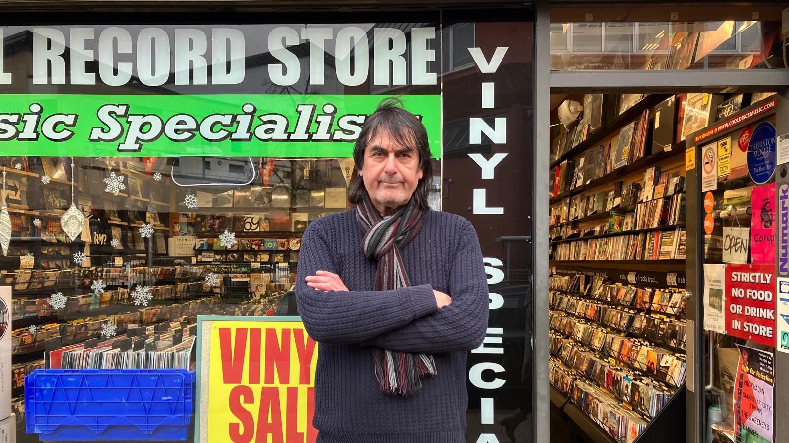 Lee Mason stands with arms crossed outside his derry city centre record shop. He is wearing a multi coloured scarf and purple jumper, he has long hair and appear to be smiling 