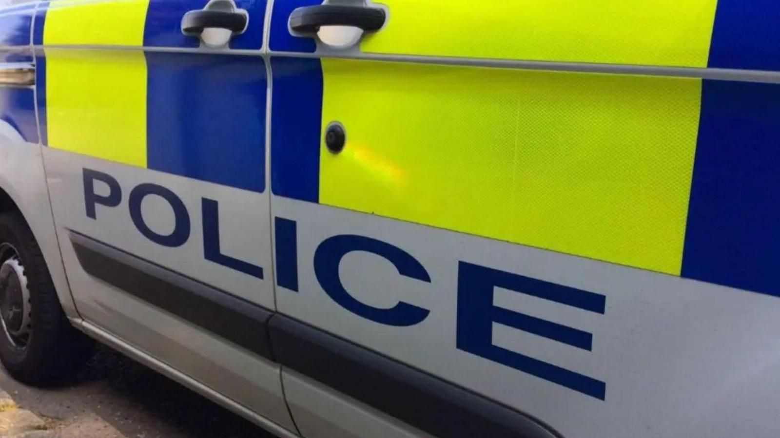 A side of a police car which is yellow and blue in colour. There is writing on the bottom of the car that says police.