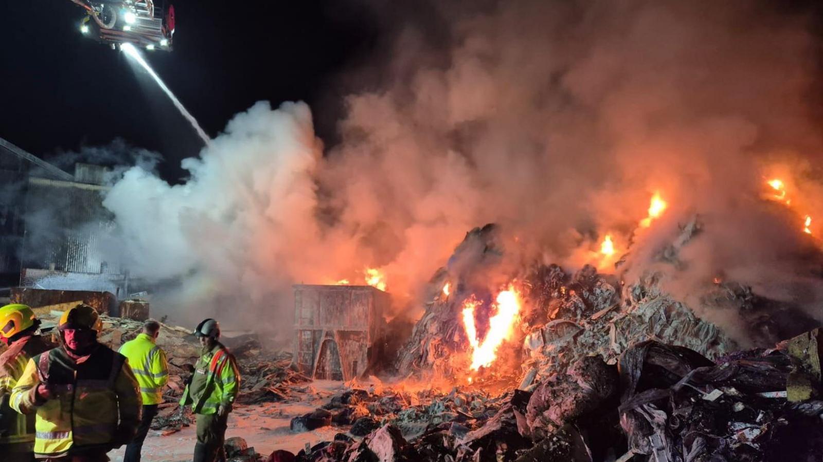 The scene of the fire at night. A small cluster of firefighters are on the left of the photo in high-vis clothing and helmets, with an aerial platform at top left squirting water on to the fire below. Flames are leaping from the mounds of rubbish or recycling with large plumes of smoke above
