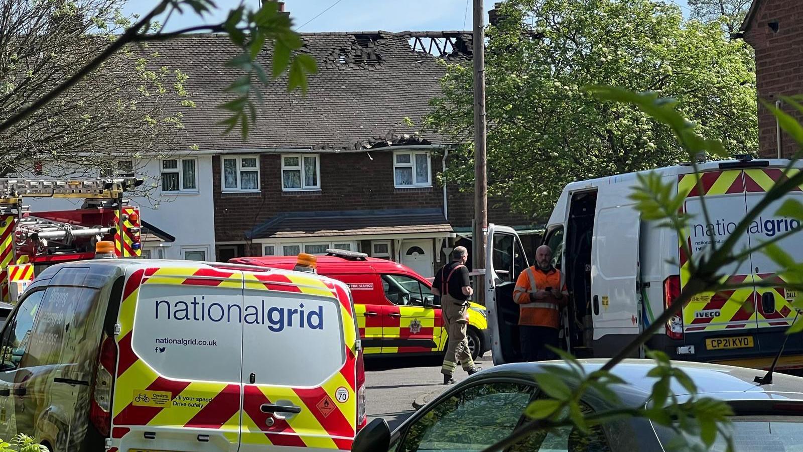 A street of terraced houses on a sunny day. One of the houses has a damaged roof with tiles missing. A fire truck can be seen in the background.