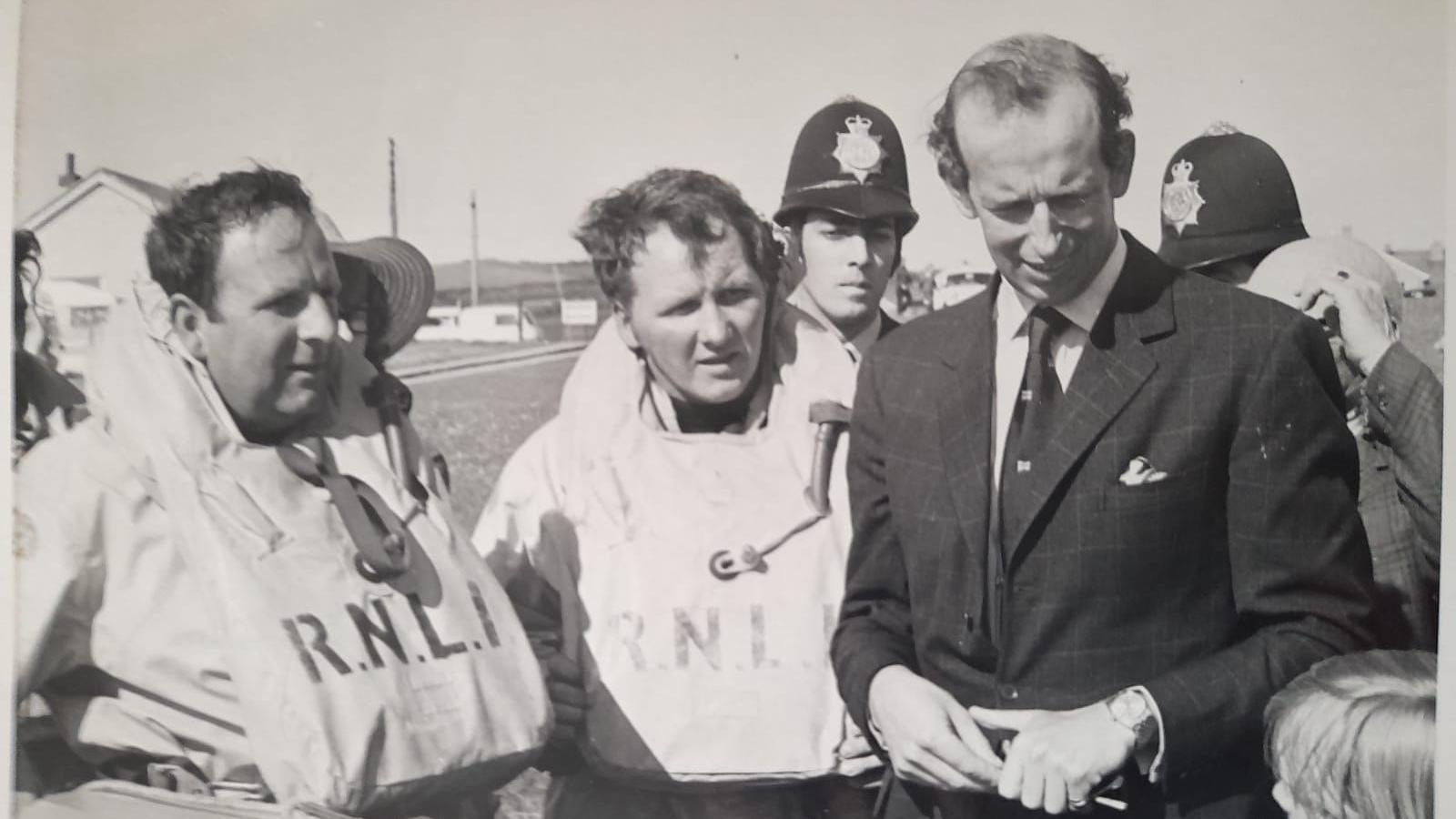 A black and white image of Bob McLaughlin and another RNLI volunteer wearing RNLI buoyancy aids, standing next to the Duke of Kent, dressed in a suit and tie, who is speaking to a child. Two police officers are behind the duke. Houses and a field can be seen in the background.