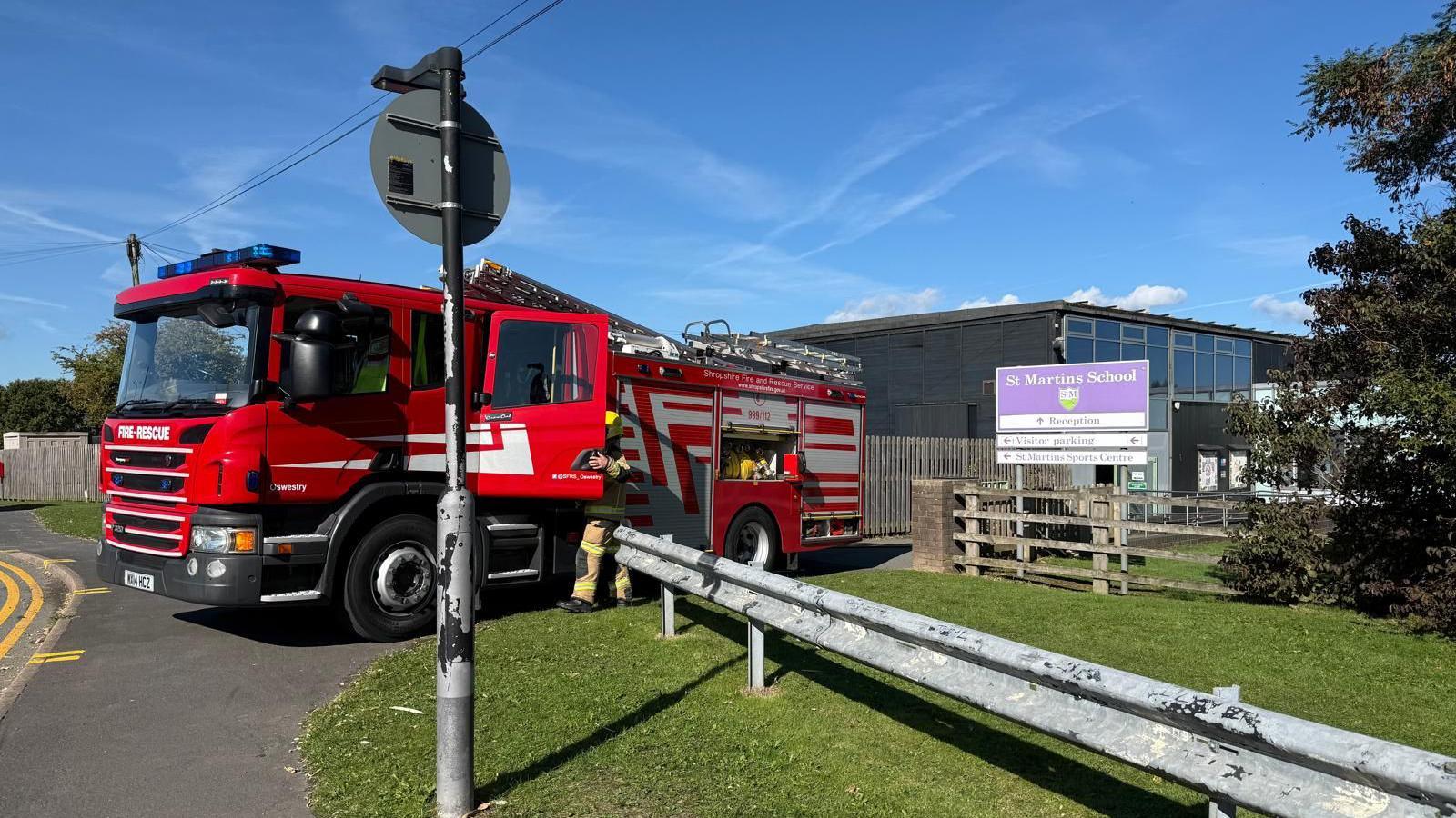 A large red and white fire engine parked outside a school building. There is a wooden gate and a purple sign that reads "St Martins School".