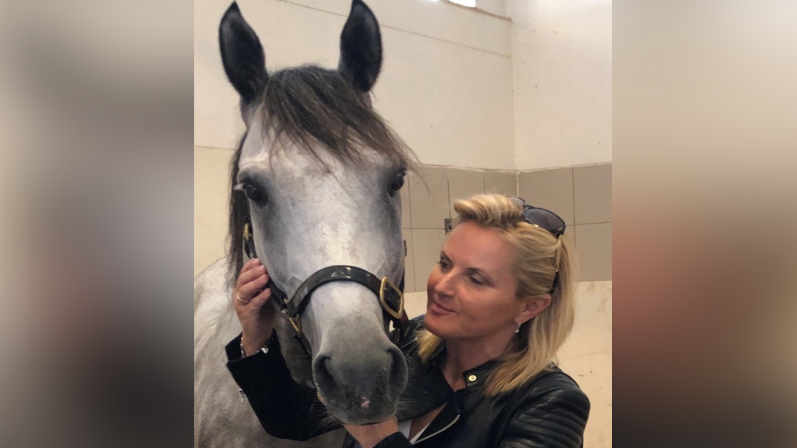 Ms Kelleway is pictured with a grey horse in a stable. She is facing the horse and holding its head in her hands while she smiles at it. She is wearing a leather jacket and sunglasses on her head. 