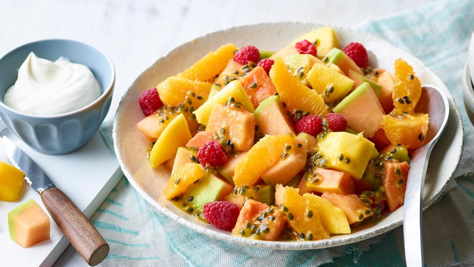 Chilled fresh fruit salad in a large serving bowl with a small dish of yoghurt next to it. There is also a chopping board in the background with two pieces of melon and a knife on it. 