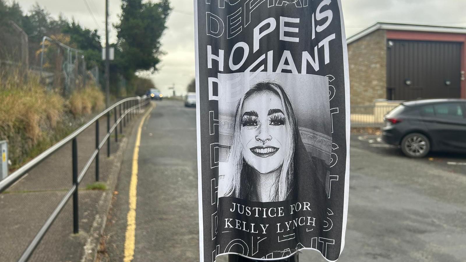 A black and white photograph of Kelly smiling is at the centre of a poster which reads 'hope is defiant' and 'Justice for Kelly Lynch'. It has been printed on a piece of A4 paper and is taped to a street light at eye level. A long black hand railing and yellow road marking line the street behind the poster, with a car driving towards the camera. 