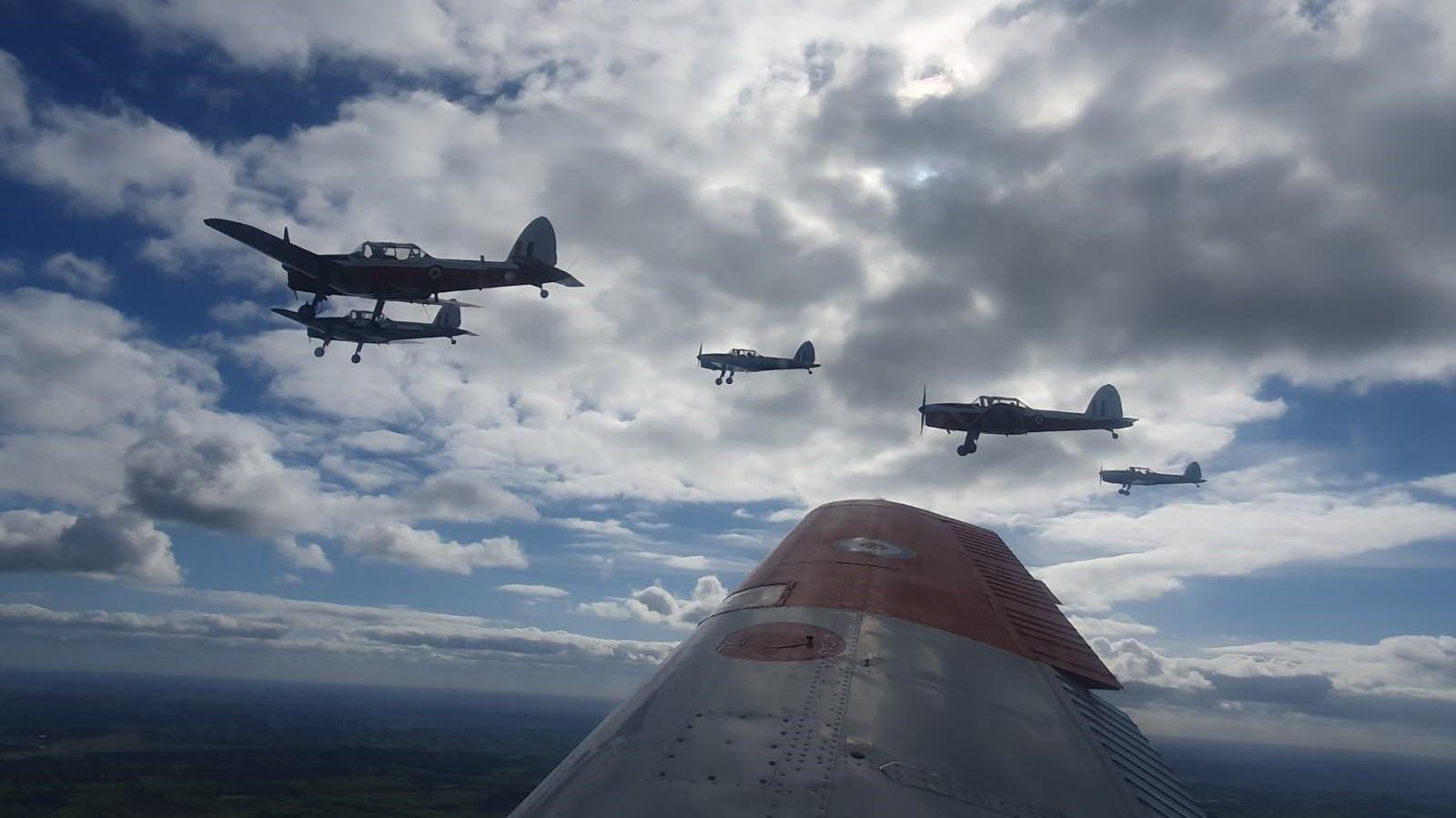 A view from the plane, and the planes fly in formation over Gloucestershire. The wing of the plane the photographer is in can be seen, and five light aircraft are in the sky. The sky is blue with lots of white clouds.