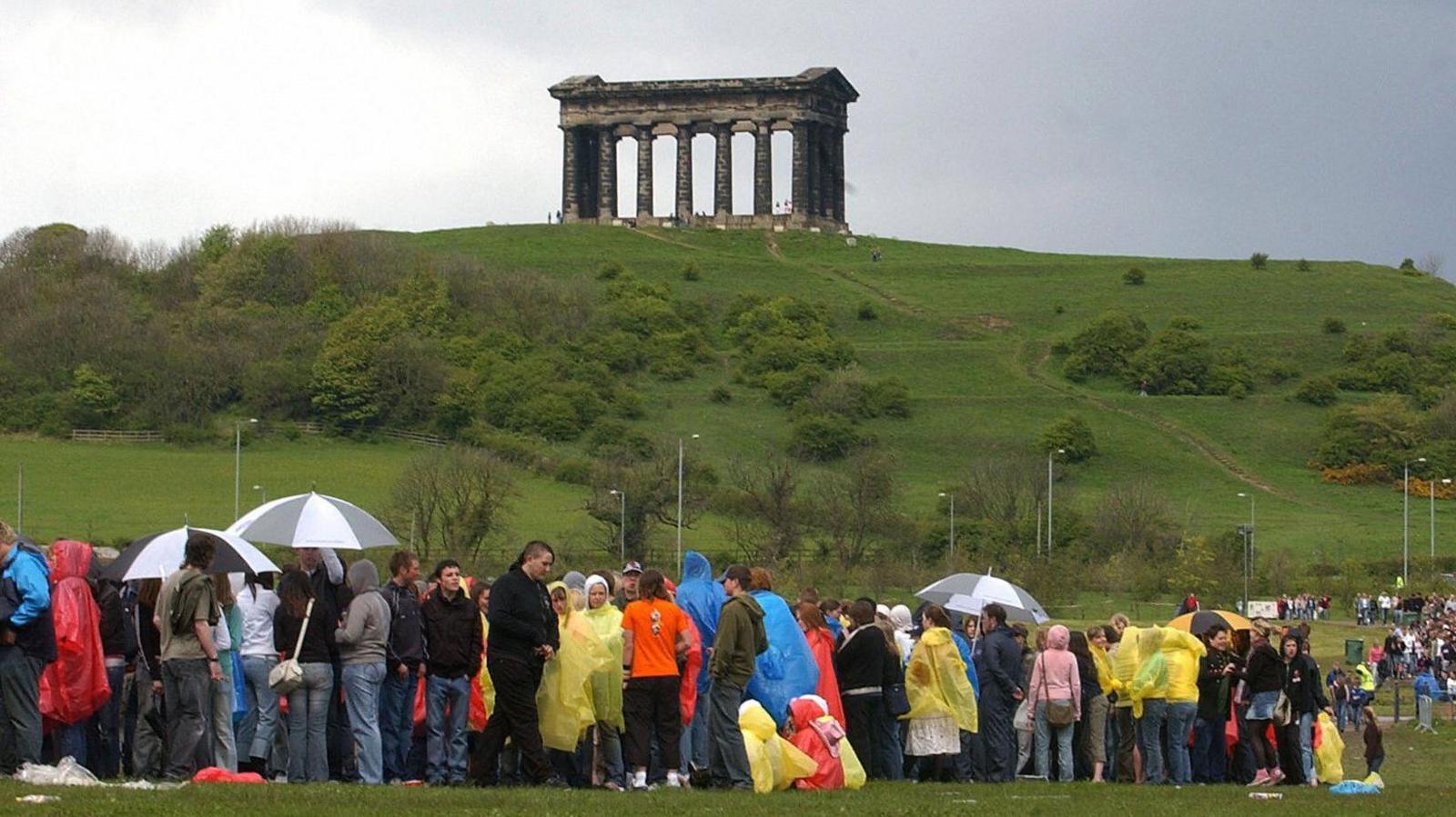 A crowd of people wearing waterproof ponchos and holding umbrellas stand in a queue in a field. On a hilltop beyond them is the Penshaw Monument, a memorial made of large stone columns.