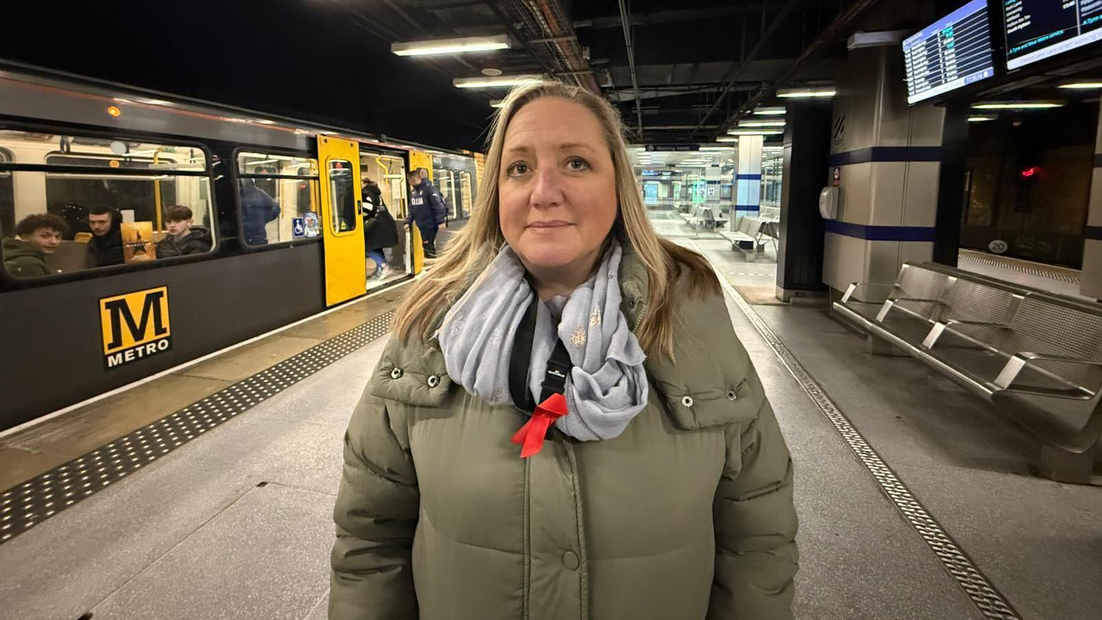 Councillor Lyndsey Leonard stands on the platform in front of a Metro train. She wears a green coat and blue scarf.