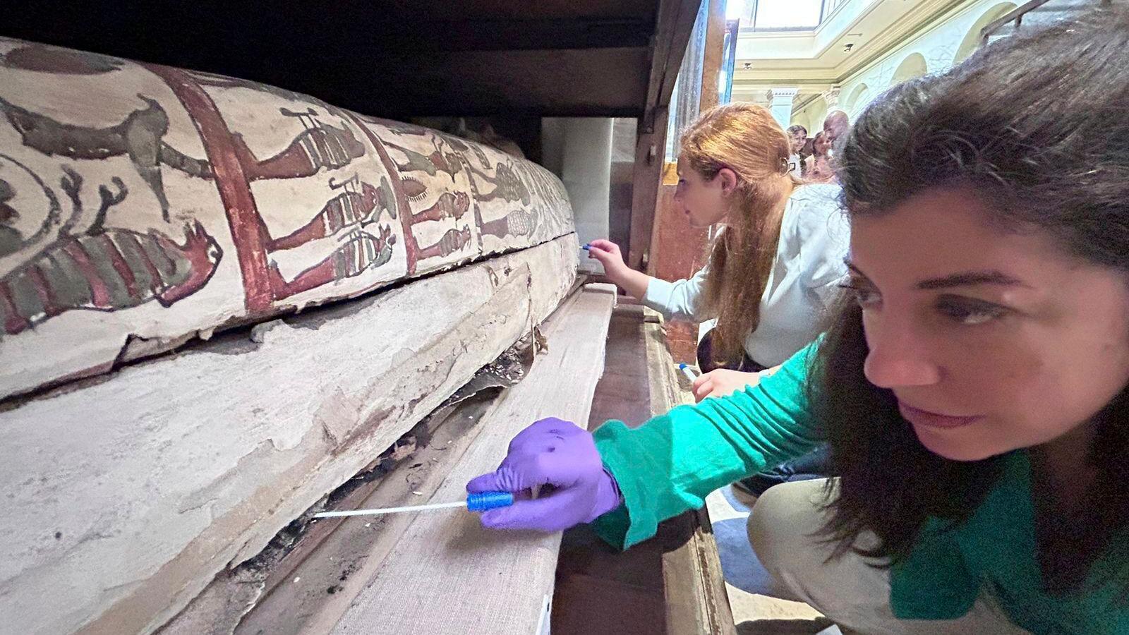 Two researches, dressed in white and wearing gloves, hold small tools up to a sarcophagus 