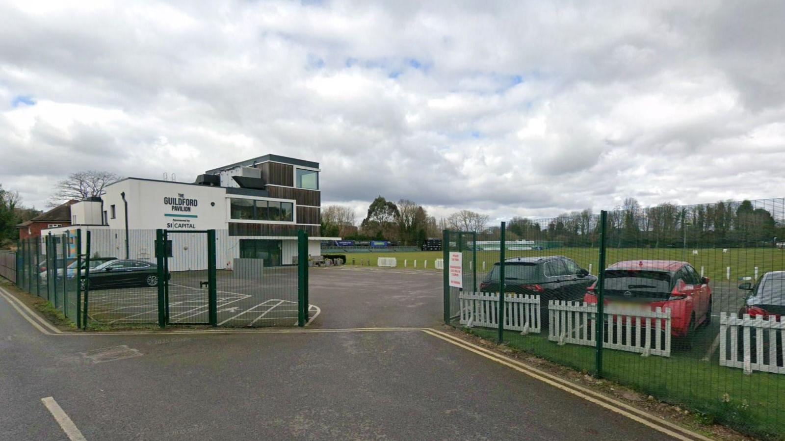 Wide shot of the main gate and fencing of Guildford Pavilion with building and green fields beyond