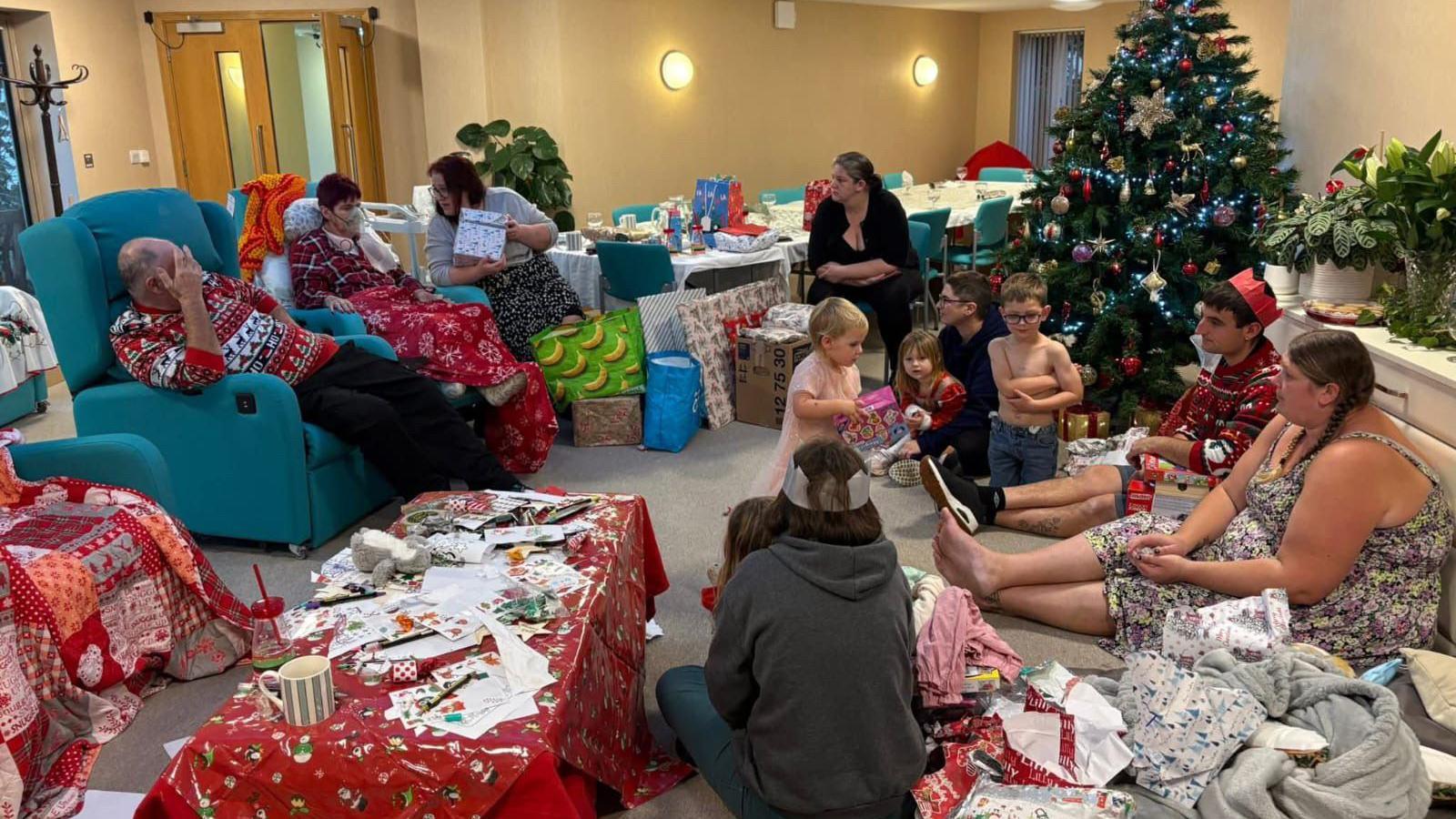 A family opening presents. There is a lot of christmas wrapping paper on the floor.