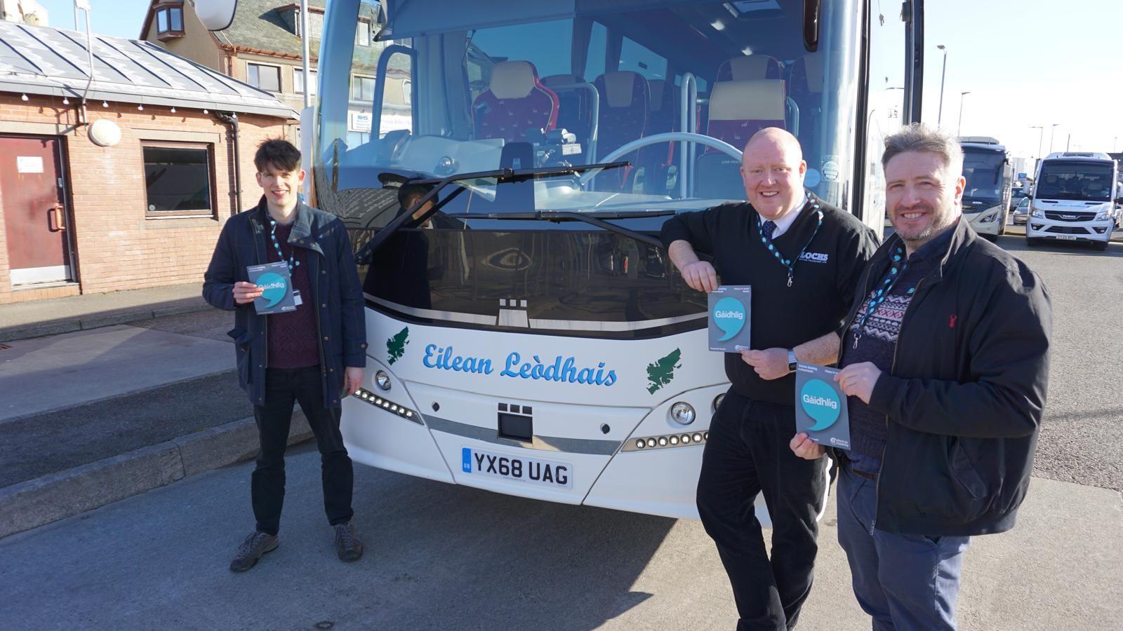 Fionnlagh Lawson bho Sgioba na Gàidhlig aig CnES, draibhear bus agus an Dr Teàrlach Wilson bho An Taigh Cèilidh air beulaibh bus agus a' cur air bhonn iomairt airson barrachd Gàidhlig a bhrosnachadh eadar draibhearan agus an luchd-siubhail