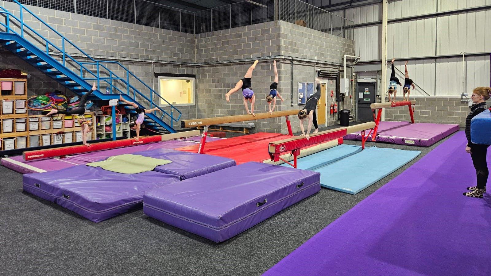 Six gymnasts are training on beams with lots of colourful mats on the floor and a coach watching on 