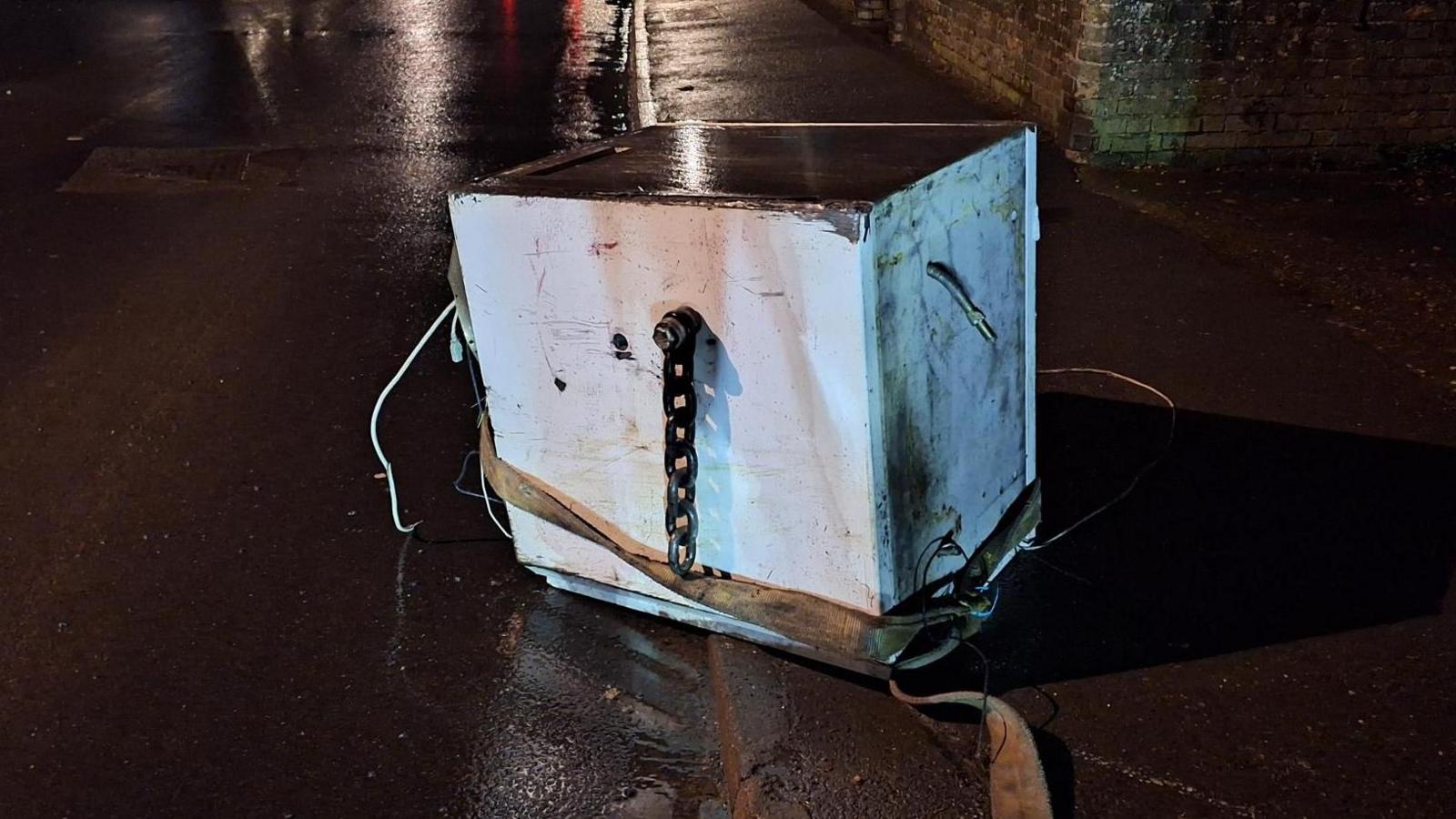 A large metal safe - from the ATM, has been dumped in the middle of a road. It has a thick chain on the front and a wide rope has been tied around it. 