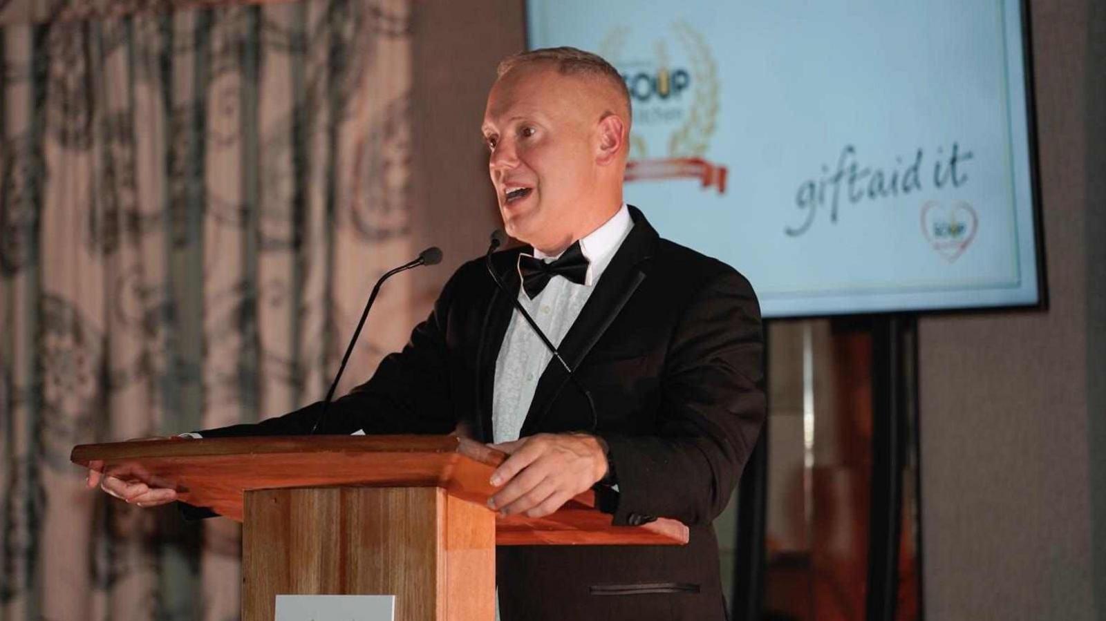 Rob Rinder smiling at the camera is wearing a black dress suit and bow tie is holding onto a podium.