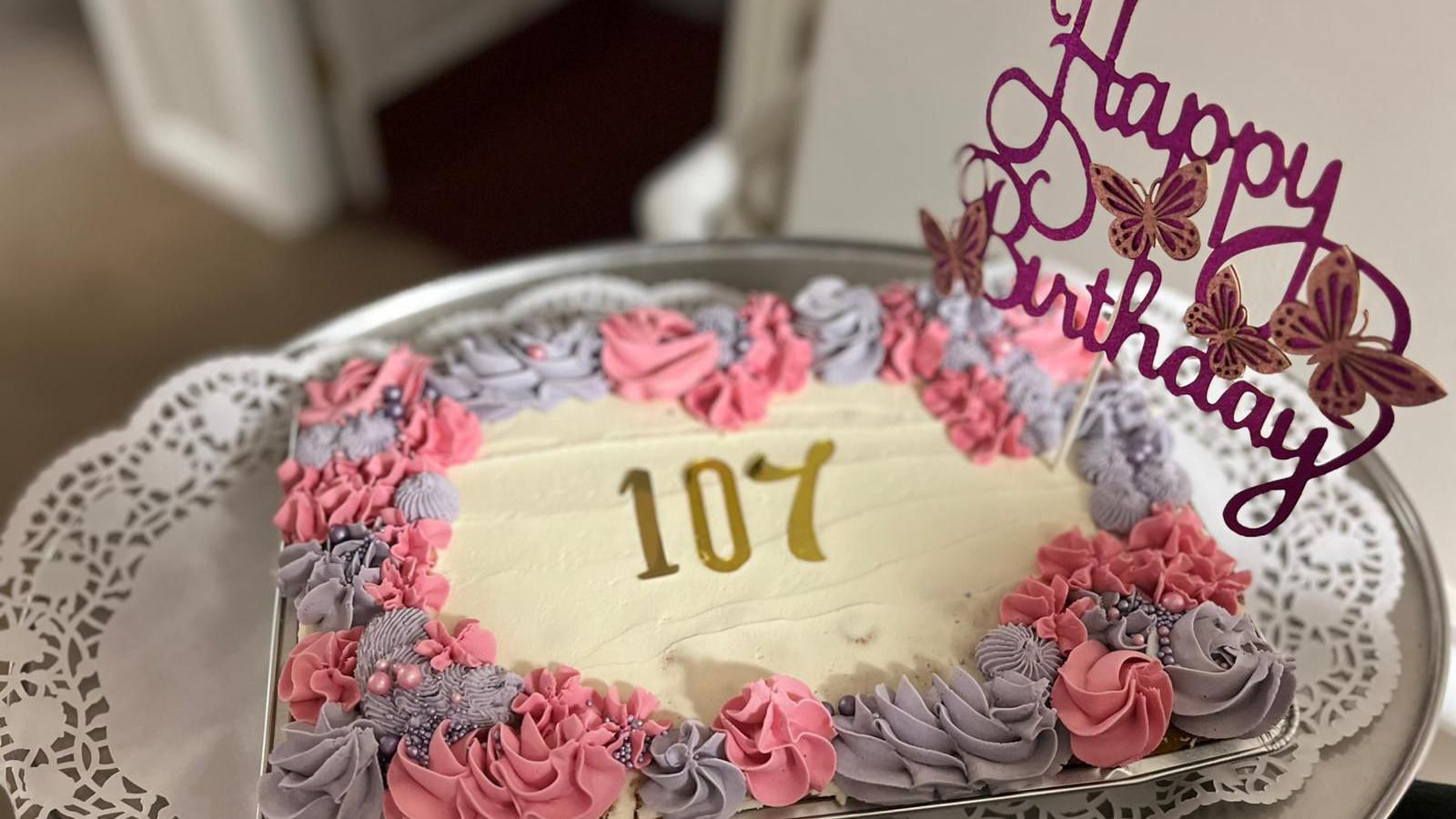 A cake with pale yellow frosting and pink and grey decoration around the edge with "107" in the middle. A topper reading "Happy Birthday" is sticking out of the cake.