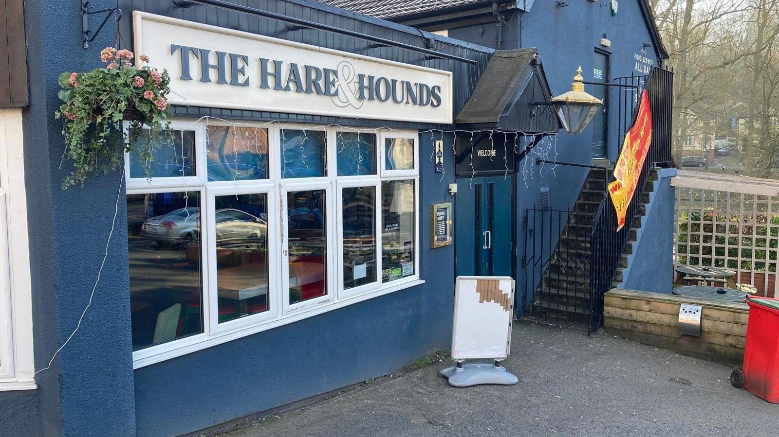 A large blue pub with the Hare and the Hounds sign on the front. There is a handing basket with pink flowers next to the sign and stairs leading up the outside of the building away from the camera. A white, blank advertising board stands outside.