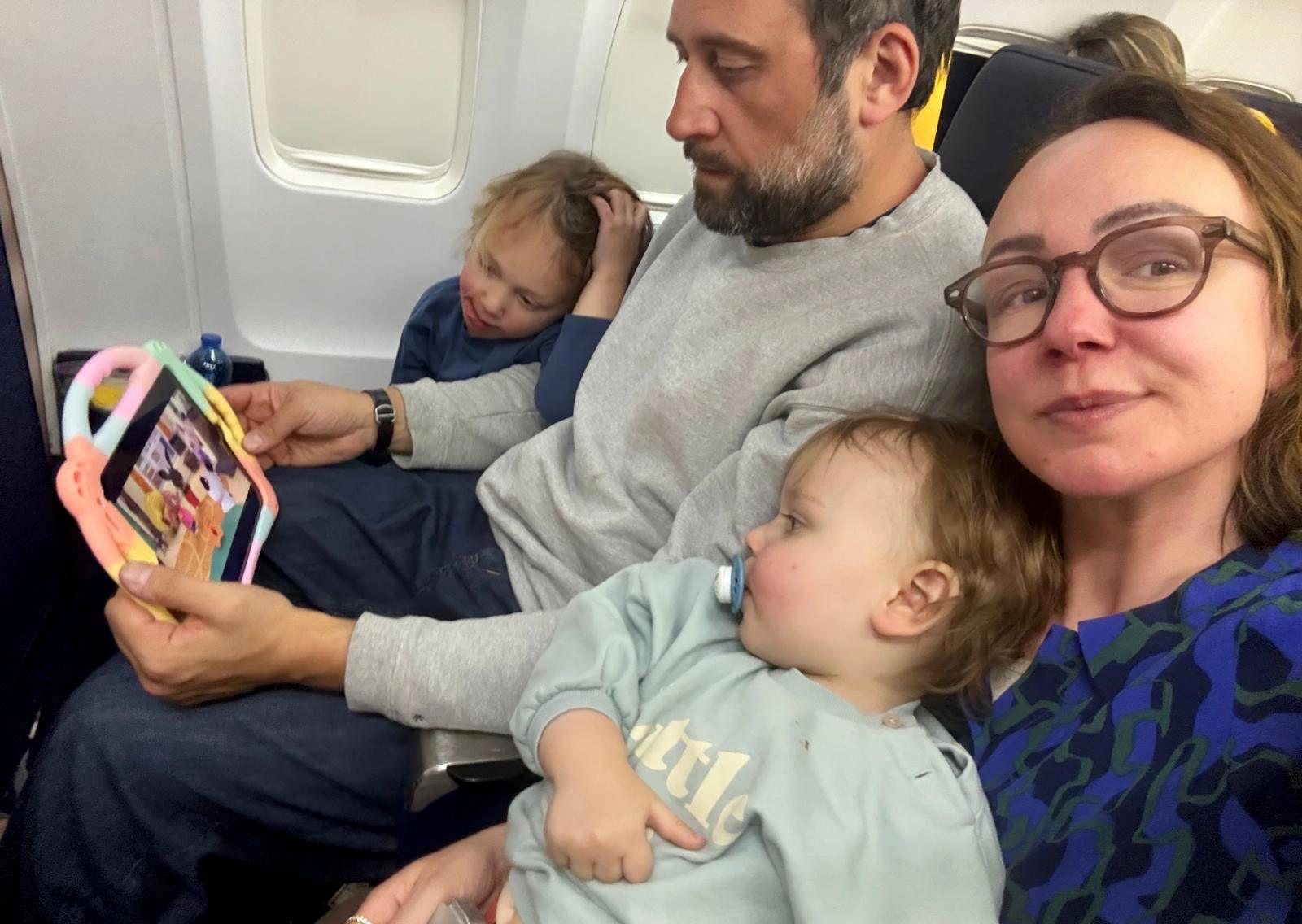 Alexis and her family sit closely together on an aeroplane, occupying two adjacent seats. She holds a tablet showing a colourful animated programme, which her two young children are watching.
