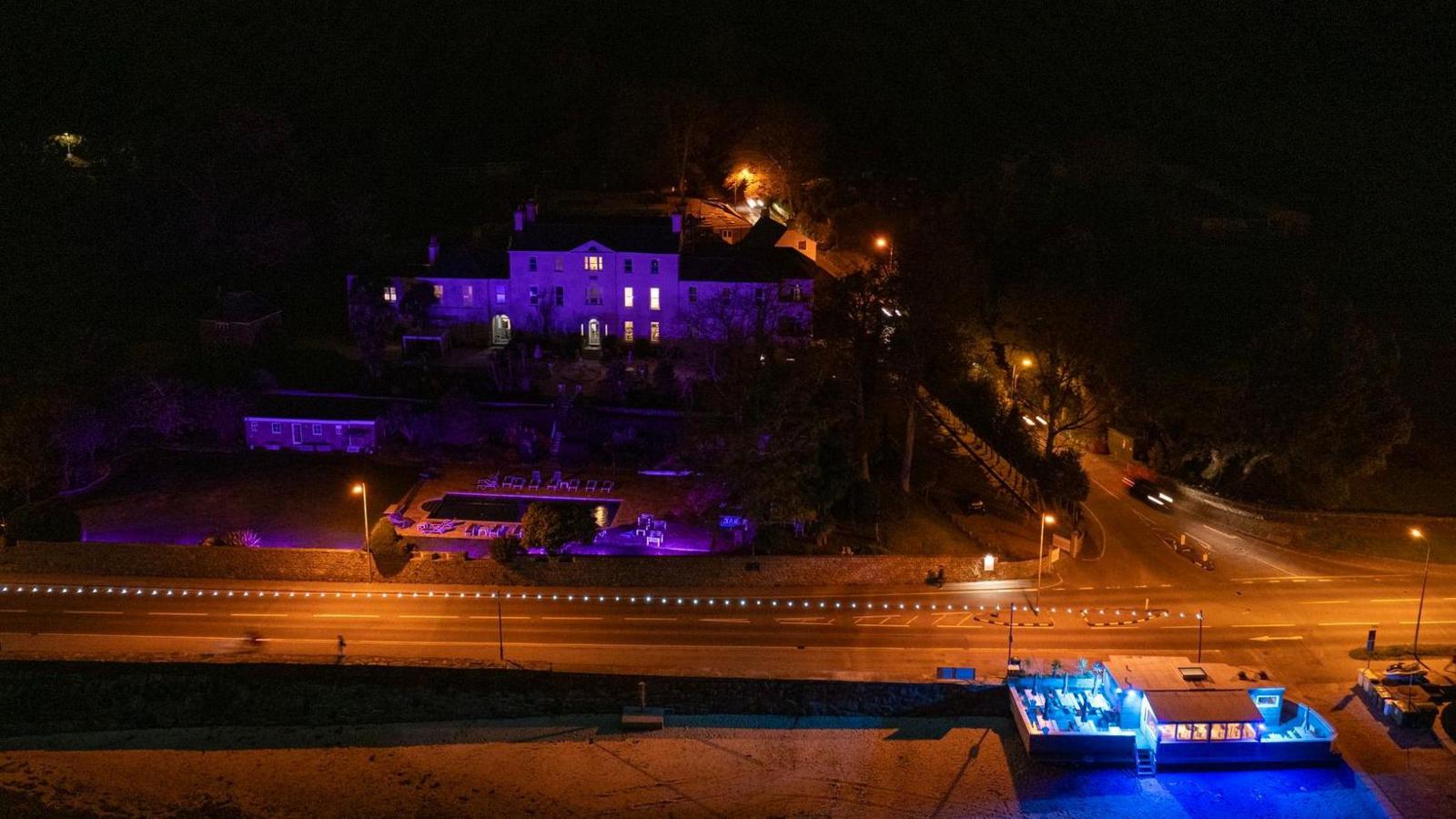 A drone shot of a sea wall, a road and a hotel lit up purple. It is night-time and streetlamps are shining a yellow light on to the road.