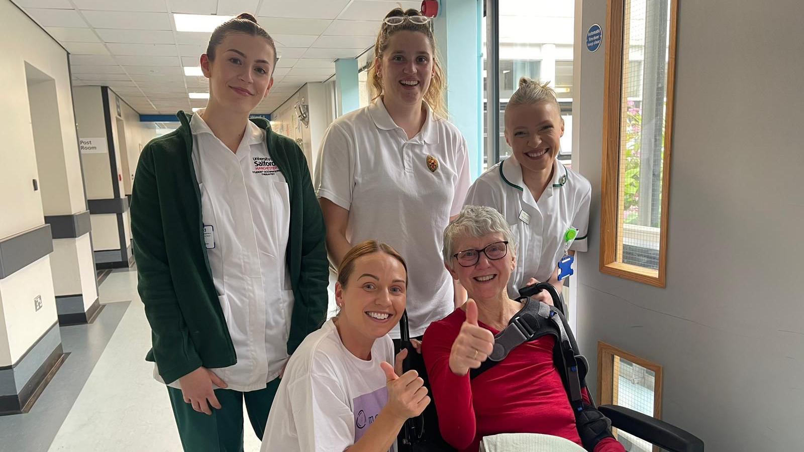 Four medical staff pose for a photograph with Maisie Farrell, a patient sat in a wheelchair in the corridor of the hospital. She is smiling with her thumb held up in the air.