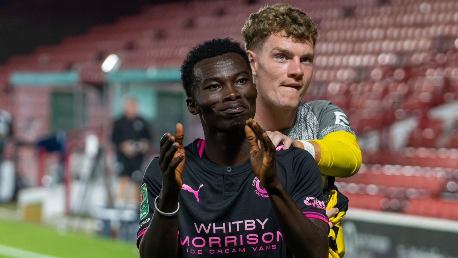 Crewe Alexandra forward Joel Ewusi applaudes the fans after becoming the club's youngest ever appearance maker and scorer. He is wearing a black shirt with pink edging and writing on it.
