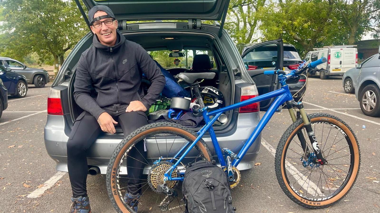 A man sits on the open pot of his car beside a blue bicycle and a black rucksack. He is wearing a dark zipped hoodie, a black rucksack, glasses and a dark baseball cap worn backwards. The boot of the car is filled with items like a sleeping bag and camping cooking equipment.