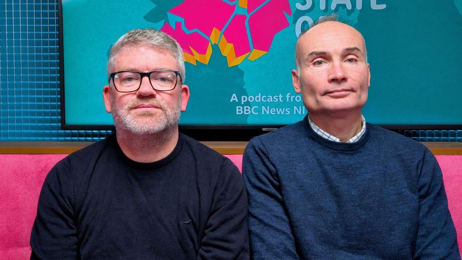 A picture of two men - Jimmy Bradley, on the left, has short grey hair and grey stubble; black-rimmed glasses; and a dark t-shirt. The man beside him, Eamon Currie, is bald and is wearing a blue jumper over a white check shirt. They are sitting in the podcast studio for The State of Us podcast. They are sitting on a pink sofa and in the background is a TV screen with The State of Us' logo - a pink rendering of Northern Ireland on a turquoise background. 