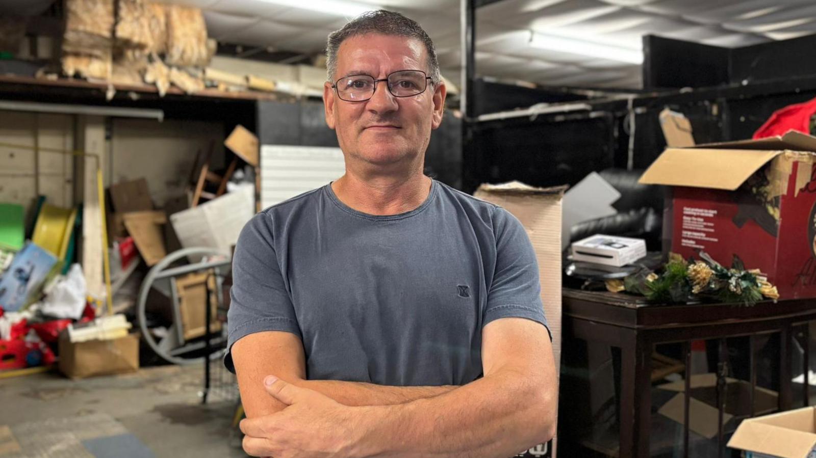 Mark Nelson has short greying dark hair and is wearing glasses standing in front of a warehouse with stock