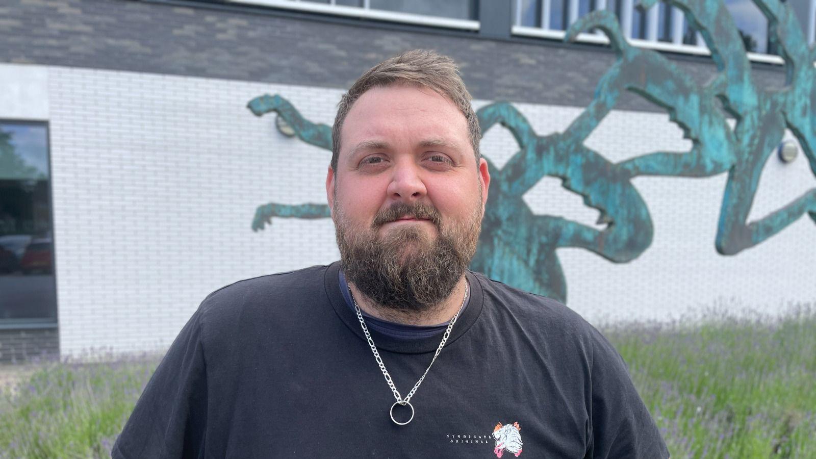 Luke is smiling at the camera, he has a dark navy t shirt on and a silver ring on a chain around his neck. He has short light brown hair and a moustache and beard.