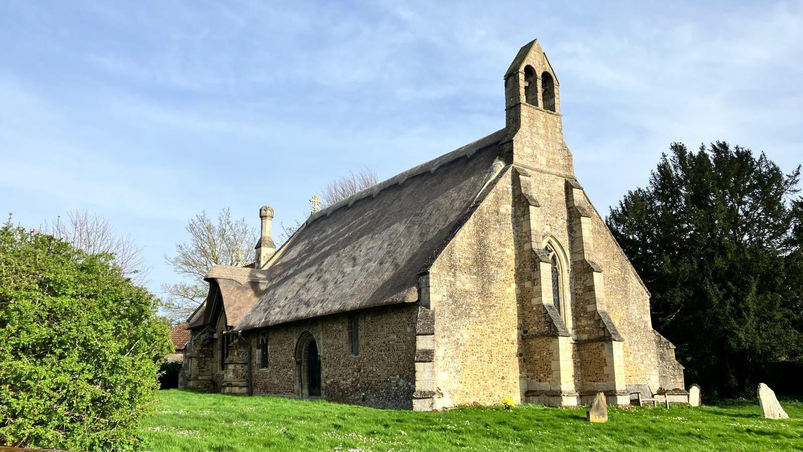 An old church is made of stone and has a thatched roof. It has a bell tower at one end with two arches. There are old gravestones on the grass outside