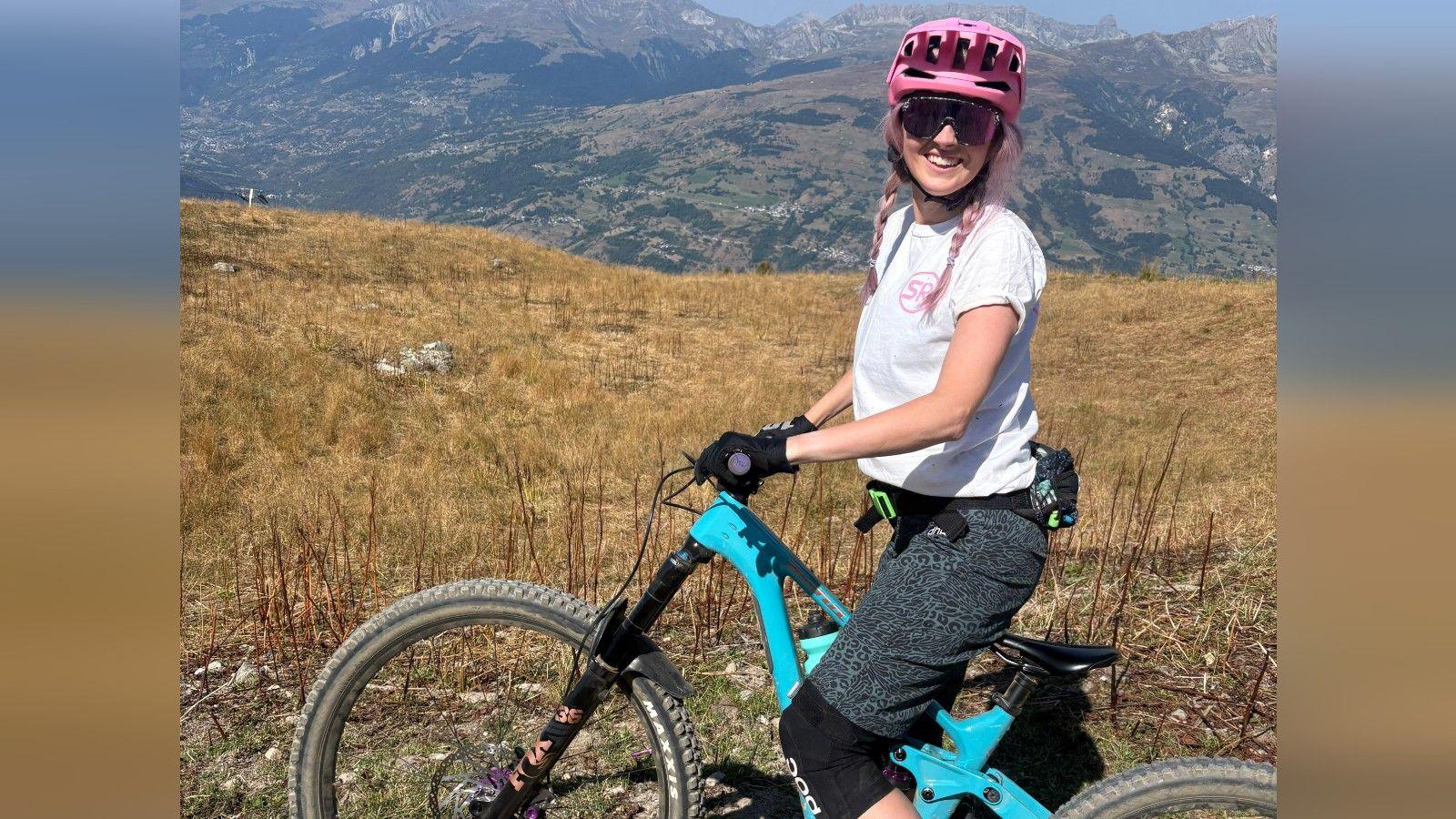 A selfie of Emma Ennis smiling while she is sat on her mountain bike, with mountains in the background. Emma is wearing a pink helmet, white t-shirt and dark shorts. She has pink hair in a two plaits. Her bike is blue, with purple handle grips.  
