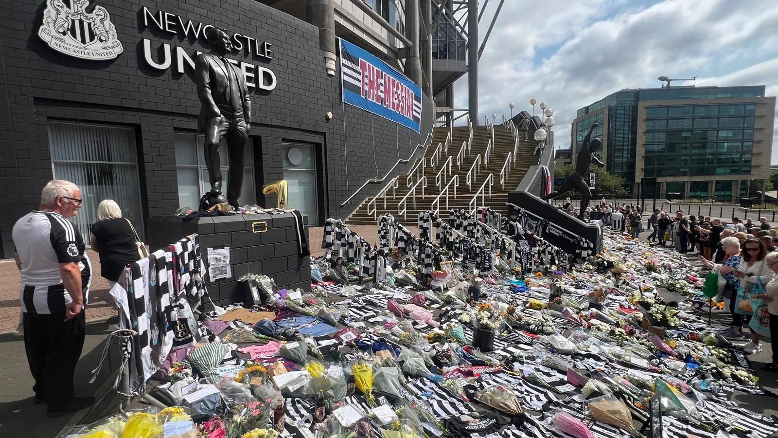 Hundreds of Newcastle United shirts, flowers, cards and other tributes on the floor outside St James' Park in Newcastle. Dozens of people are lined up looking at the tributes to Kevin Keegan. There is a blue and white banner hanging from the side of the stadium, between the Newcastle United sign and the steps, with the words The Messiah, in tribute to Kevin Keegan.