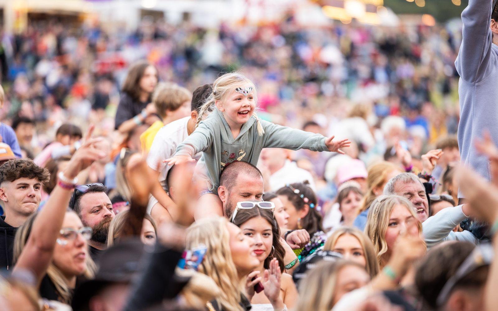 Young child dances on a parents shoulders surrounded by a smiling crowd
