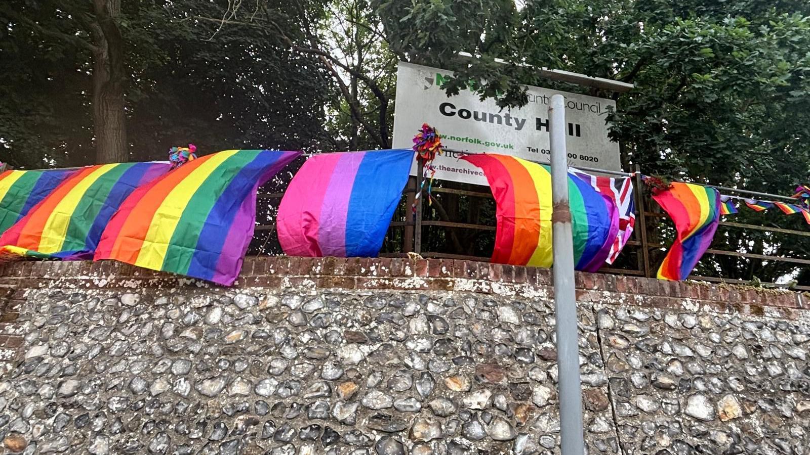 Flags being flown from outside the entrance to Norfolk County Hall in Norwich. The rainbow striped flags are fixed to metal railings above a stone wall. A white sign reads COUNTY HALL, and there are trees behind it.
